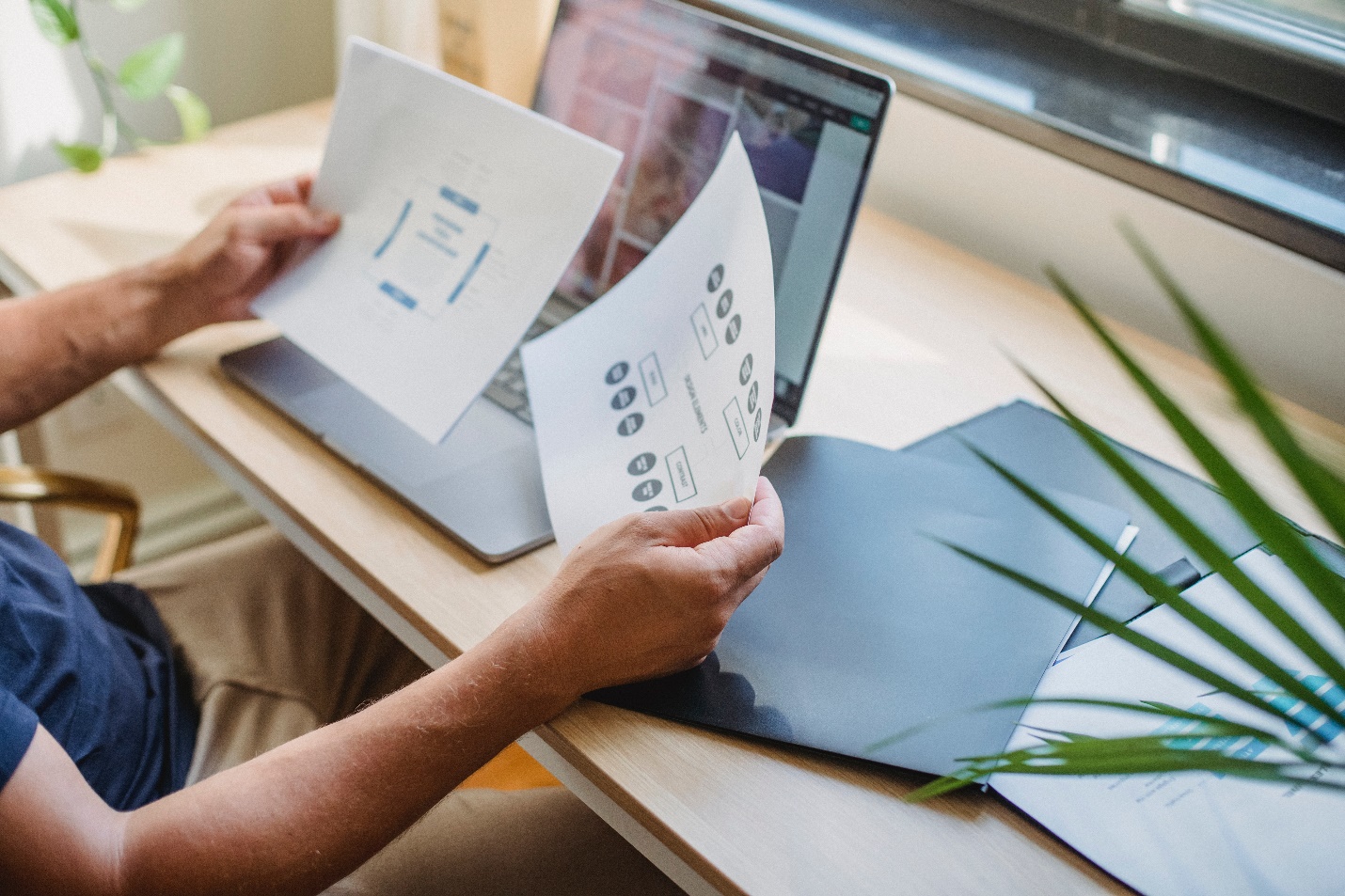 A man sits at a desk holding two sheets of paper with a website development plan and an open laptop on his desk